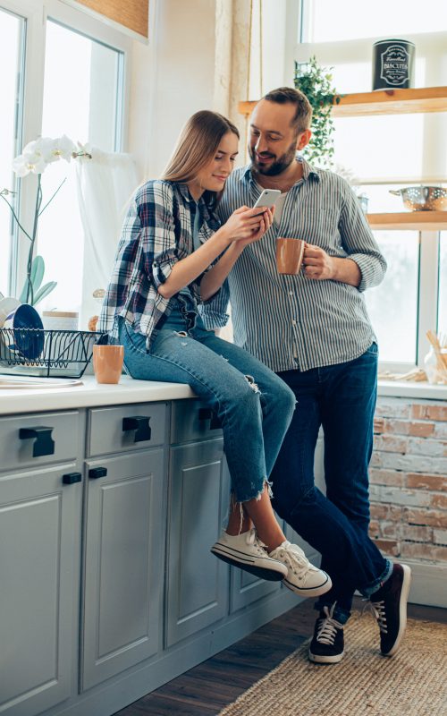 Pretty lady sitting in the kitchen by the sink and using smartphone while being with boyfriend